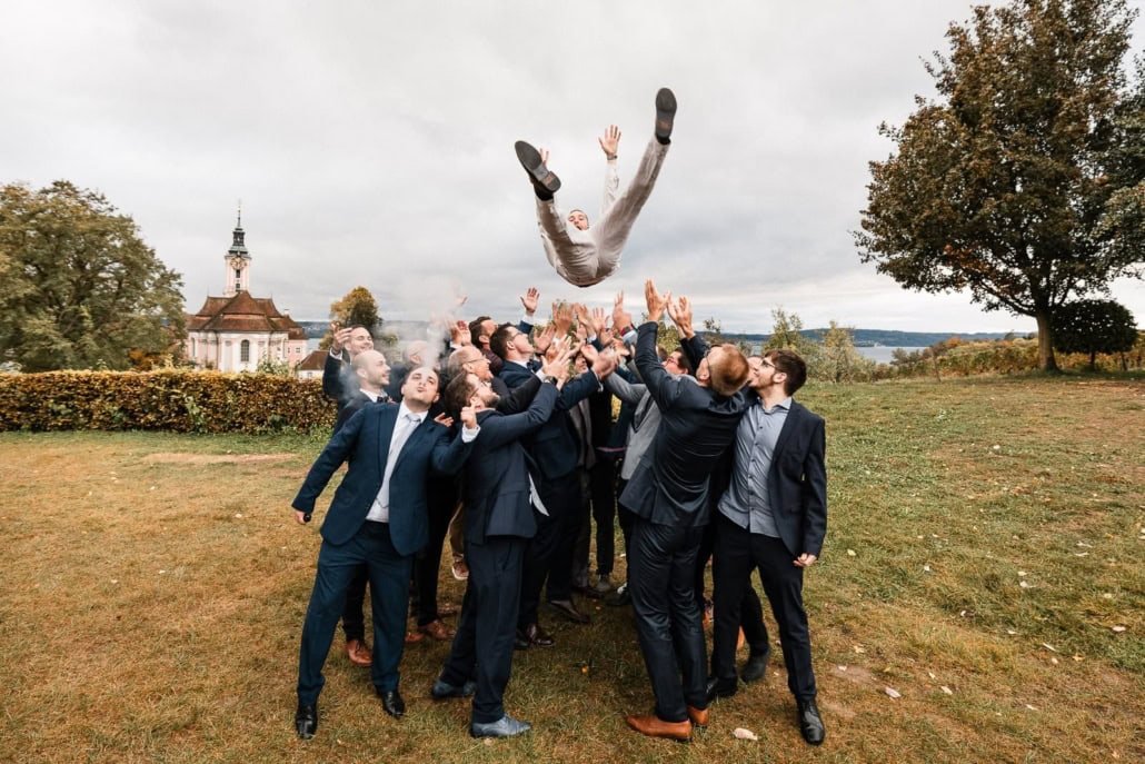 Eine Gruppe von Menschen in Anzügen wirft einen Mann lächelnd und jubelnd in die Luft. Im Hintergrund sind Bäume, Gras und eine Kirche unter einem bewölkten Himmel zu sehen.