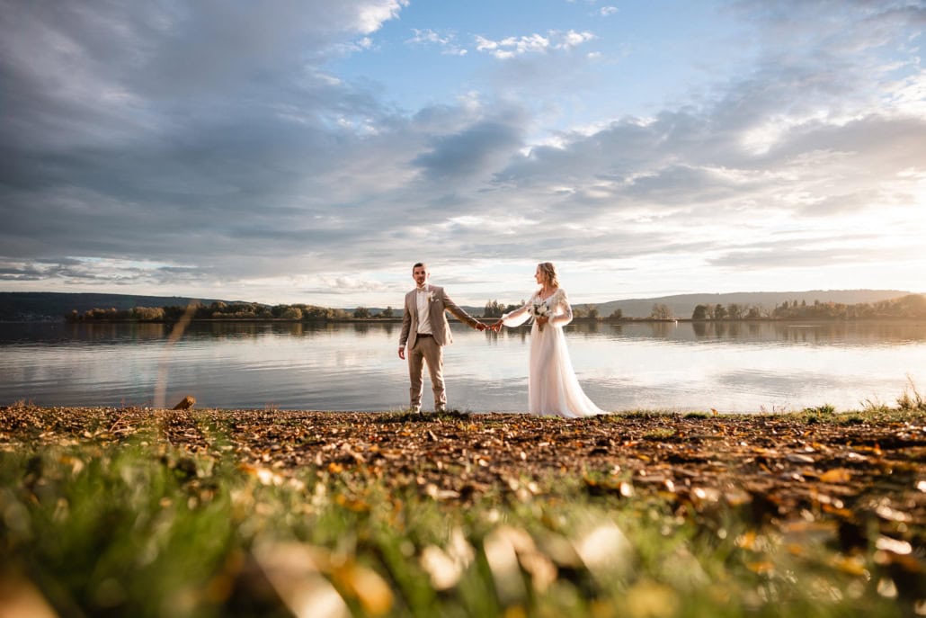 Eine Braut und ein Bräutigam stehen Hand in Hand an einem grasbewachsenen Seeufer bei Sonnenuntergang, mit ruhigem Wasser und einem dramatischen Wolkenhimmel im Hintergrund - perfekt eingefangen von ihrem Hochzeitsfotografen.