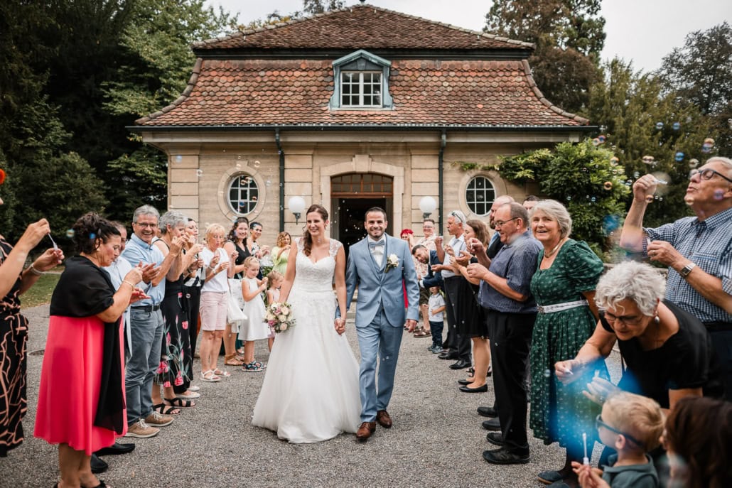 Ein frisch verheiratetes Paar geht lächelnd Hand in Hand, während die Hochzeitsgäste auf beiden Seiten stehen, Seifenblasen blasen und klatschen. Aufgenommen von einem Hochzeitsfotografen, stehen sie vor einem charmanten Gebäude mit Ziegeldach und umgeben von Grün.