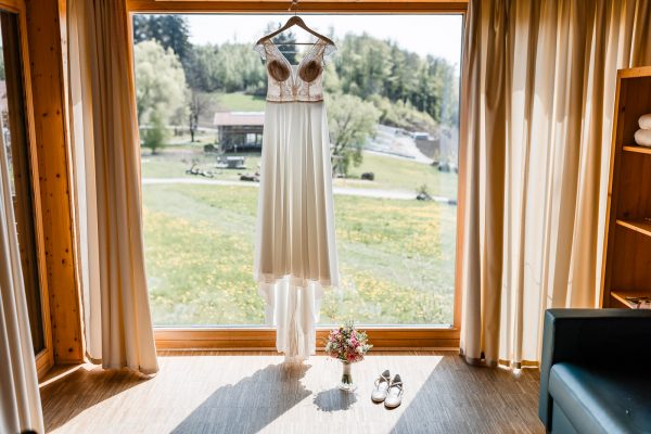 Brautkleid, Brautstrauß und Brautschuhe vor Panoramafenster auf der Gutsalm Harlachberg
